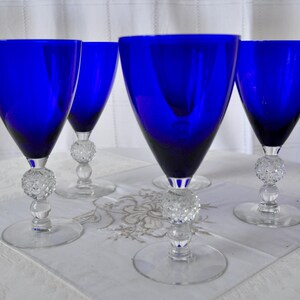 May include: A set of four cobalt blue glass goblets with clear stems. Each goblet features a deep blue bowl and a clear glass stem with a textured, spherical knob detail above a circular base. The glasses are arranged in a group on a white surface.