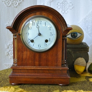 May include: A wooden antique clock with a white face and black numerals. The clock has a dark brown finish and a curved top. The clock's hands point to nearly 12 o'clock. The clock is a decorative item.