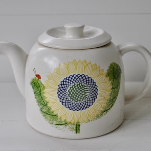May include: White ceramic teapot with a sunflower design. The sunflower is yellow with a blue and green center. A small red ladybug is on a green leaf.