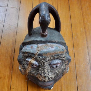 Puede incluir: Una m&aacute;scara tribal antigua y desgastada con una superficie oscura y texturizada. La m&aacute;scara presenta un asa arqueada prominente con un dise&ntilde;o de serpiente en la parte superior. La cara tiene ojos detallados, un &aacute;rea de boca perforada y una superficie agrietada.