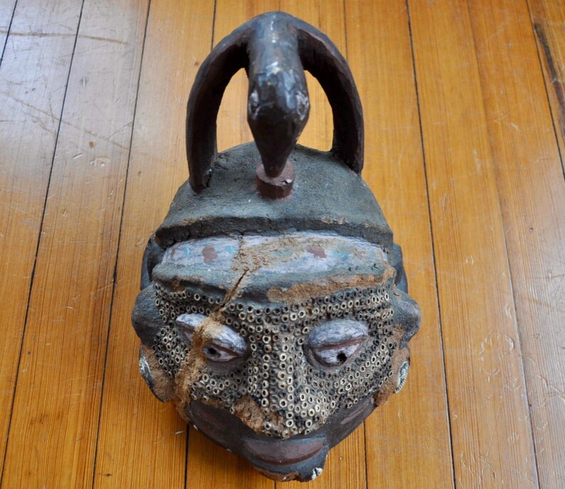Puede incluir: Una m&aacute;scara tribal antigua y desgastada con una superficie oscura y texturizada. La m&aacute;scara presenta un asa arqueada prominente con un dise&ntilde;o de serpiente en la parte superior. La cara tiene ojos detallados, un &aacute;rea de boca perforada y una superficie agrietada.
