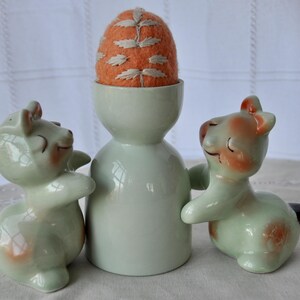 Puede incluir: Un huevero de cer&aacute;mica vintage con un coj&iacute;n alfiletero en forma de huevo de fieltro naranja. Dos figuras de animales de cer&aacute;mica a juego flanquean el huevero. Las figuras son de color verde claro con detalles en melocot&oacute;n y negro.