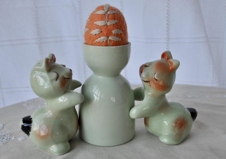 Puede incluir: Un huevero de cer&aacute;mica vintage con un coj&iacute;n alfiletero en forma de huevo de fieltro naranja. Dos figuras de animales de cer&aacute;mica a juego flanquean el huevero. Las figuras son de color verde claro con detalles en melocot&oacute;n y negro.