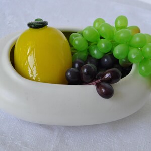 May include: A smooth, off-white, oval-shaped ceramic bowl containing a glossy yellow glass lemon with a green stem and a cluster of artificial green and dark purple grapes. A small, dark, rectangular label is attached to the side of the bowl.