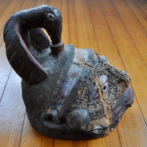 Puede incluir: Una escultura artesanal de color oscuro con un asa en forma de p&aacute;jaro. La escultura tiene una superficie texturizada con un patr&oacute;n de peque&ntilde;os agujeros circulares en un lado. El dise&ntilde;o sugiere una pieza de arte antiguo o tribal.