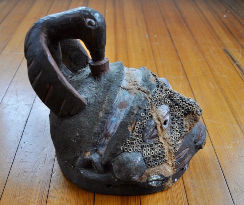 Puede incluir: Una escultura artesanal de color oscuro con un asa en forma de p&aacute;jaro. La escultura tiene una superficie texturizada con un patr&oacute;n de peque&ntilde;os agujeros circulares en un lado. El dise&ntilde;o sugiere una pieza de arte antiguo o tribal.