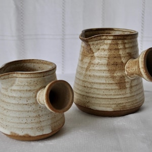 May include: Two ceramic pitchers with a cream-colored base and brown speckles. The pitchers have a spout and a cylindrical handle. The larger pitcher is approximately 6 inches tall, and the smaller pitcher is about 4 inches tall. The pitchers are on a white surface.