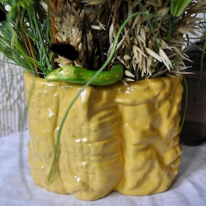 May include: A yellow ceramic vase with a textured surface, filled with dried wheat, green grass, and black accents. A green ceramic accent sits on the rim. The vase is on a white surface.