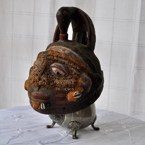 Puede incluir: Escultura de madera tallada de una cabeza humana con una peque&ntilde;a figura de animal en la parte superior. La cabeza es de color marr&oacute;n oscuro con detalles en naranja y beige, y tiene una base de metal con tres patas ornamentadas.