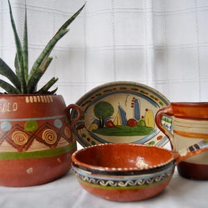 May include: A terracotta planter with the word MEXICO painted on it, containing a green plant, alongside an oval dish with a landscape painting, a small terracotta bowl with a handle, and a cylindrical mug with brown and cream horizontal stripes.