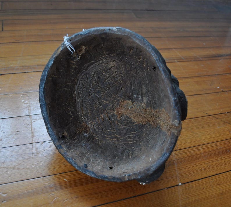 Puede incluir: Una m&aacute;scara de madera tallada de color marr&oacute;n oscuro con forma redondeada. El interior muestra marcas de tallado visibles y una textura rugosa. La m&aacute;scara tiene un borde ligeramente elevado y parece hecha a mano.