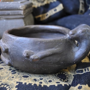 May include: A dark gray, handmade ceramic bowl with a unique, organic shape. The bowl features a raised, textured design and a small, bird-like handle. Perfect for holding small items or as a decorative piece. The bowl is approximately 6 inches in diameter.