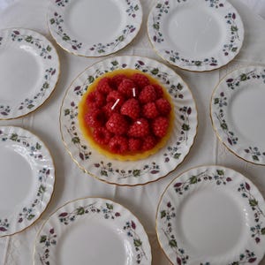 May include: A set of seven white ceramic plates with scalloped edges and a delicate floral pattern featuring green leaves and small blue and purple flowers, with one plate holding a round yellow candle topped with realistic red raspberry shapes.