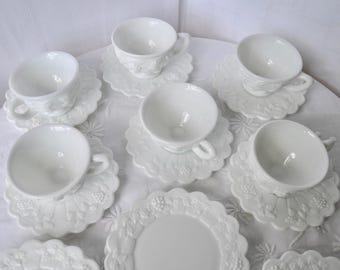May include: A collection of white ceramic teacups and saucers. The cups and saucers have a detailed embossed floral design. The set includes multiple cups and saucers, and a plate, all arranged on a white surface.
