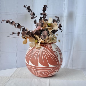 May include: A spherical terracotta vase featuring white etched geometric and fan-shaped patterns, filled with a dried floral arrangement consisting of brown eucalyptus stems and pale yellow flowers with prominent green centers.
