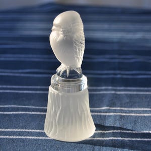 May include: A clear and frosted glass bell featuring a sculpted owl perched on top. The handle is shaped like a detailed owl with textured feathers, while the bell body has a vertical, bark-like pattern. The glass transitions from clear at the top to a matte, frosted finish on the lower section.