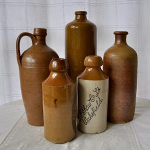 May include: A collection of five vintage stoneware bottles and jugs in various shades of brown and tan. One cylindrical bottle features the black printed text J. Shaw & Co. Ltd. Wakefield. The vessels have narrow necks and varied textures.