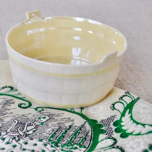 May include: A white ceramic bowl with a yellow rim and a green and white pattern. The bowl is sitting on a green and white patterned fabric.