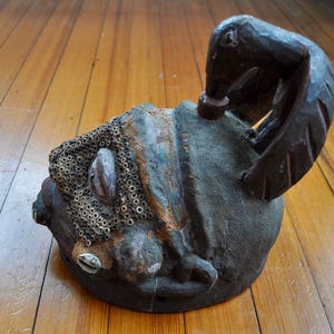 Puede incluir: Una m&aacute;scara de madera tallada a mano, desgastada, con una figura de ave en la parte superior. La m&aacute;scara presenta una superficie texturizada con una mezcla de tonos marrones, azules y beige. Detalles incluyen un acento de concha y un patr&oacute;n de peque&ntilde;os elementos circulares.