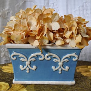 May include: A blue rectangular ceramic planter with white scrollwork holds dried, beige-colored flowers. The planter sits on a table with a floral pattern, creating a vintage aesthetic. The flowers add a touch of natural beauty.