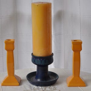 May include: A tall yellow pillar candle sits in a dark blue ceramic candle holder, flanked by two matching yellow ceramic candlesticks. The candle holder features a dark blue base with a decorative pattern of teal leaf-like shapes.