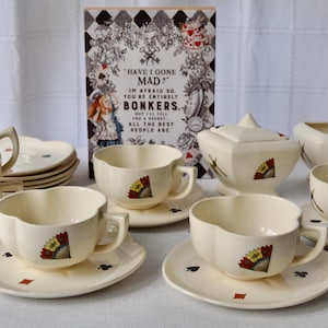 May include: Cream-colored teacups and saucers with a fan design and playing card symbols. A sugar bowl and a stack of plates are also visible. A decorative sign with the text "HAVE I GONE MAD?" is in the background. The tea set is likely vintage.