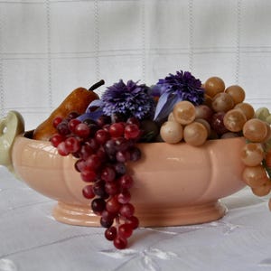 May include: A peach-colored ceramic fruit bowl filled with artificial grapes, a pear, and purple flowers. The bowl has a decorative handle on each side and sits on a white tablecloth.