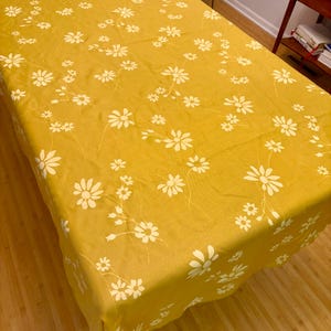 May include: A rectangular tablecloth in a vibrant mustard yellow color featuring a repeating pattern of white daisy-like flowers with thin stems and leaves. The fabric drapes over the edges of a table, displaying a consistent floral print throughout.