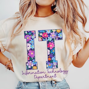 May include: Ivory-colored t-shirt with the letters "IT" in a floral design. The text "Information Technology Department" is printed below the letters. The shirt is paired with blue jeans.
