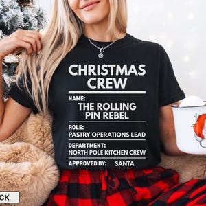 May include: A black short-sleeved t-shirt featuring white text that reads CHRISTMAS CREW, NAME: THE ROLLING PIN REBEL, ROLE: PASTRY OPERATIONS LEAD, DEPARTMENT: NORTH POLE KITCHEN CREW, and APPROVED BY: SANTA, with a white mug showing a Santa graphic.
