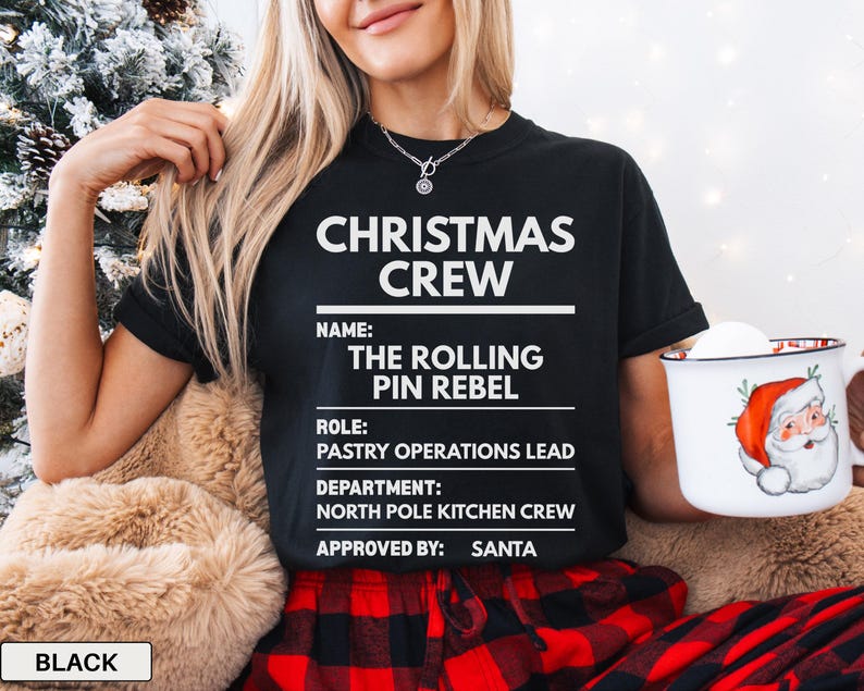 May include: A black short-sleeved t-shirt featuring white text that reads CHRISTMAS CREW, NAME: THE ROLLING PIN REBEL, ROLE: PASTRY OPERATIONS LEAD, DEPARTMENT: NORTH POLE KITCHEN CREW, and APPROVED BY: SANTA, with a white mug showing a Santa graphic.