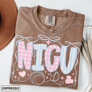 A folded espresso Comfort Colors T-shirt with "NICU" spelled out in large, patchwork-style letters. The "N" and "C" are soft pink gingham, while the "I" and "U" are light blue and white vertical stripes. Surrounding the letters are pastel pink ribbons, looping thread, sparkles, a pink gingham heart, and pink ABC building blocks. The style is sweet, nurturing, and whimsical, perfect for NICU nurses and staff.