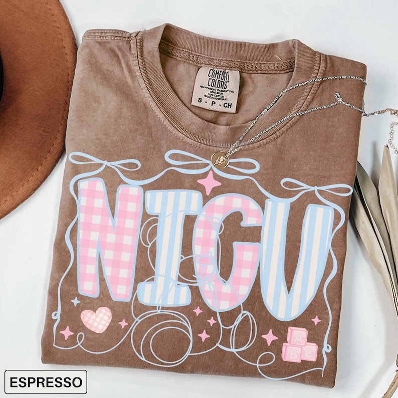 A folded espresso Comfort Colors T-shirt with "NICU" spelled out in large, patchwork-style letters. The "N" and "C" are soft pink gingham, while the "I" and "U" are light blue and white vertical stripes. Surrounding the letters are pastel pink ribbons, looping thread, sparkles, a pink gingham heart, and pink ABC building blocks. The style is sweet, nurturing, and whimsical, perfect for NICU nurses and staff.