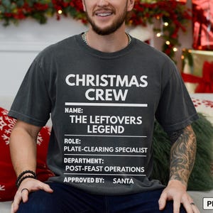May include: A charcoal gray short-sleeve t-shirt featuring white text that reads CHRISTMAS CREW, NAME: THE LEFTOVERS LEGEND, ROLE: PLATE-CLEARING SPECIALIST, DEPARTMENT: POST-FEAST OPERATIONS, and APPROVED BY: SANTA, arranged in a structured list format.