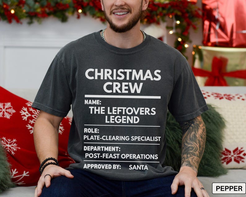 May include: A charcoal gray short-sleeve t-shirt featuring white text that reads CHRISTMAS CREW, NAME: THE LEFTOVERS LEGEND, ROLE: PLATE-CLEARING SPECIALIST, DEPARTMENT: POST-FEAST OPERATIONS, and APPROVED BY: SANTA, arranged in a structured list format.