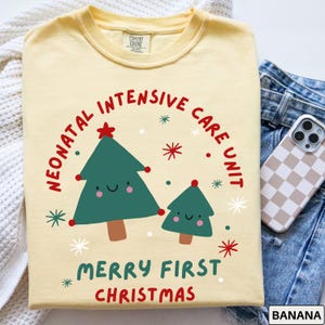 Comfort Colors banana yellow tshirt featuring a festive NICU design. The graphic shows two cute Christmas trees, one large and one small, surrounded by colorful red and green starbursts and snowflakes. Bold curved text reads “NEONATAL INTENSIVE CARE UNIT” above, with “MERRY FIRST CHRISTMAS” below in playful red and green lettering, creating a cheerful holiday look for NICU staff and families.