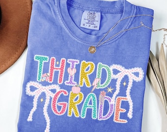 May include: A blue t-shirt with the words "THIRD GRADE" in colorful, playful lettering. The design includes hearts, bows, and a lightning bolt. The shirt is labeled "FLO BLUE" at the bottom.