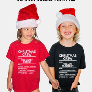 May include: A red t-shirt and a black t-shirt featuring white text that reads COMFORT COLORS YOUTH TEE, CHRISTMAS CREW, NAME: THE SMART COOKIE, ROLE: HOLIDAY STRATEGY MANAGER, DEPARTMENT: CHRISTMAS BRAIN TRUST, APPROVED BY: SANTA, and THE MINI MUNCHKIN.