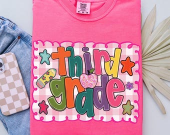 May include: A bright pink t-shirt with the words "Third Grade" in colorful, playful lettering. The design includes a pencil, apple, stars, and flowers on a pink and white gingham background. The shirt is folded on top of blue jeans.