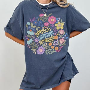 Puede incluir: Camiseta Comfort Colors 1717 azul denim con un dise&ntilde;o floral y las palabras "Speech Language Pathologist". El dise&ntilde;o incluye flores rosas, amarillas, azules y moradas. La camiseta es de corte holgado.