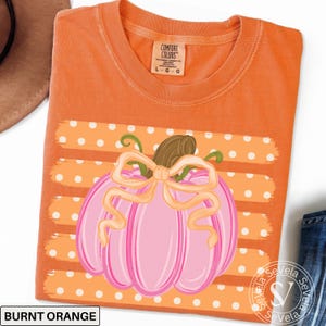 May include: A burnt orange t-shirt featuring a graphic of a pink pumpkin with a light orange bow. The pumpkin is centered over horizontal stripes with white polka dots. The inner neck label reads Comfort Colors, 100% cotton, made in Honduras.