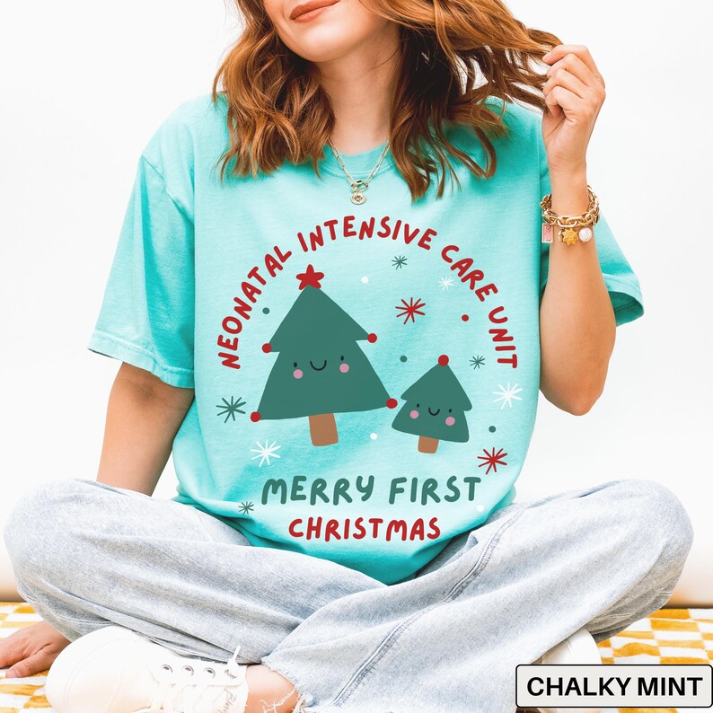 May include: A light teal t-shirt with a graphic featuring two cartoon Christmas trees and the text "Neonatal Intensive Care Unit Merry First Christmas." The design includes red stars and the text "Chalky Mint."