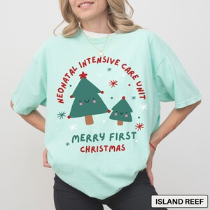 May include: A light green t-shirt with a festive design. The shirt features a graphic with two cartoon Christmas trees, snowflakes, and the text "Neonatal Intensive Care Unit Merry First Christmas" in red. The shirt is a comfortable, casual style.