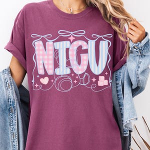 May include: A maroon oversized t-shirt with a graphic design that says "NICU" in pink and blue checkered and striped letters. The graphic also includes hearts, stars, and pacifiers.