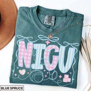 A folded blue spruce Comfort Colors T-shirt with "NICU" spelled out in large, patchwork-style letters. The "N" and "C" are soft pink gingham, while the "I" and "U" are light blue and white vertical stripes. Surrounding the letters are pastel pink ribbons, looping thread, sparkles, a pink gingham heart, and pink ABC building blocks. The style is sweet, nurturing, and whimsical, perfect for NICU nurses and staff.