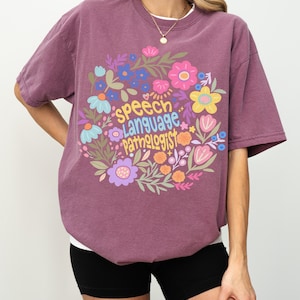 Puede incluir: Camiseta color bayas con dise&ntilde;o floral y la frase "Speech Language Pathologist". La camiseta de manga corta tiene cuello redondo. La modelo lleva un pantal&oacute;n corto negro. La camiseta es de corte holgado.