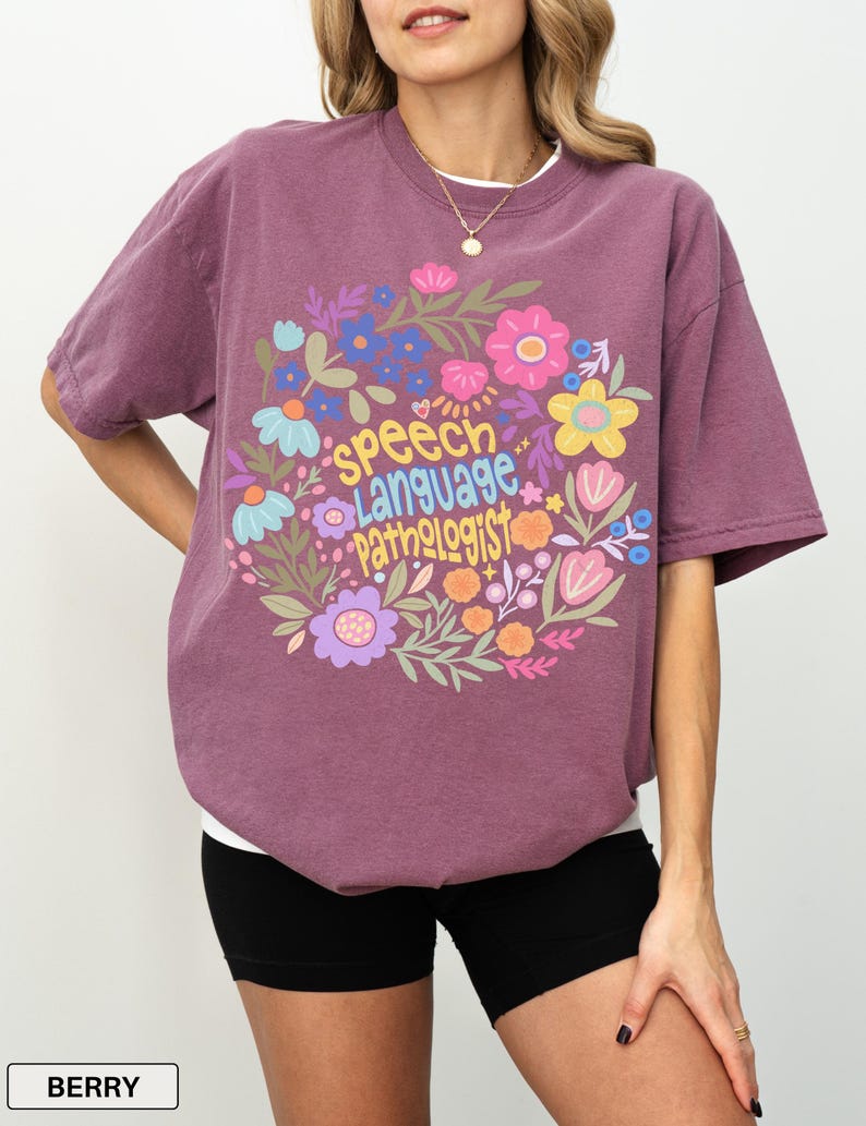 Puede incluir: Camiseta color bayas con dise&ntilde;o floral y la frase "Speech Language Pathologist". La camiseta de manga corta tiene cuello redondo. La modelo lleva un pantal&oacute;n corto negro. La camiseta es de corte holgado.