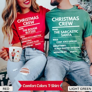May include: A red t-shirt with white text reading CHRISTMAS CREW, THE BOUGIE BLITZEN, SLEIGH STYLE DIRECTOR, HOLIDAY GLAM, and SANTA, and a green t-shirt with white text reading CHRISTMAS CREW, THE SARCASTIC SANTA, CHIEF JOKE OFFICER, SLEIGH BANTER DIVISION, and SANTA.