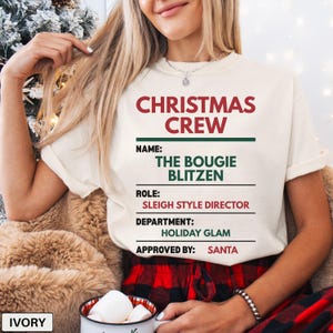 May include: An ivory t-shirt featuring red and green text that reads CHRISTMAS CREW, NAME: THE BOUGIE BLITZEN, ROLE: SLEIGH STYLE DIRECTOR, DEPARTMENT: HOLIDAY GLAM, and APPROVED BY: SANTA. The design is printed on the front of the short-sleeved shirt.
