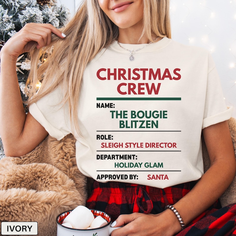 May include: An ivory t-shirt featuring red and green text that reads CHRISTMAS CREW, NAME: THE BOUGIE BLITZEN, ROLE: SLEIGH STYLE DIRECTOR, DEPARTMENT: HOLIDAY GLAM, and APPROVED BY: SANTA. The design is printed on the front of the short-sleeved shirt.